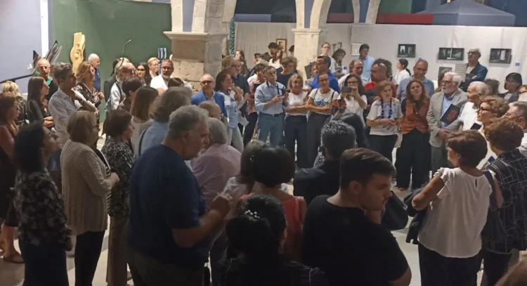 Pubblico in piedi tra archi in pietra e quadri appesi a pannelli bianchi alla mostra del Collettivo BAI a Comiso