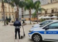 Agente della Polizia Locale di spalle accanto a un monopattino elettrico in una piazza di Comiso, vicino a un’auto di servizio