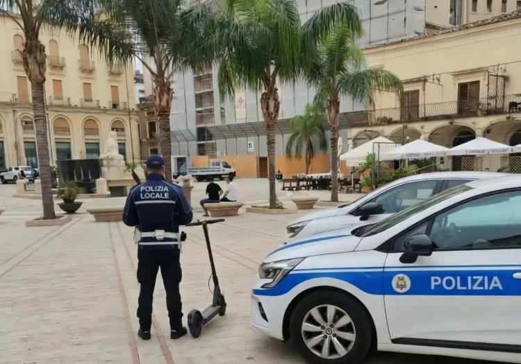 Agente della Polizia Locale di spalle accanto a un monopattino elettrico in una piazza di Comiso, vicino a un’auto di servizio