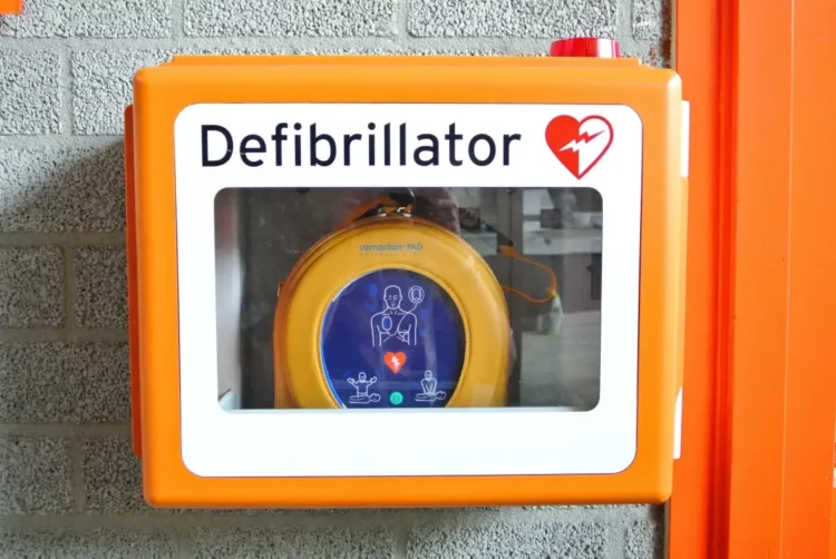 Defibrillatore semiautomatico giallo in una teca arancione fissata a un muro di mattoni grigi, con il simbolo del cuore rosso