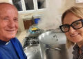 Don Beniamino Sacco sorride in una cucina di Vittoria accanto a grandi pentole fumanti, insieme a una donna con occhiali scuri