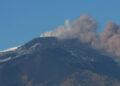 Pennacchio vulcanico grigio e rosato si alza dalla cima innevata dell’Etna e si allunga verso destra sotto un cielo azzurro