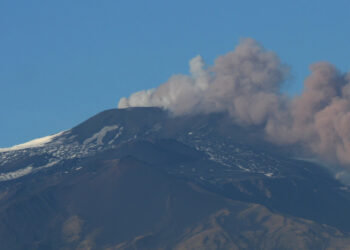 Pennacchio vulcanico grigio e rosato si alza dalla cima innevata dell’Etna e si allunga verso destra sotto un cielo azzurro