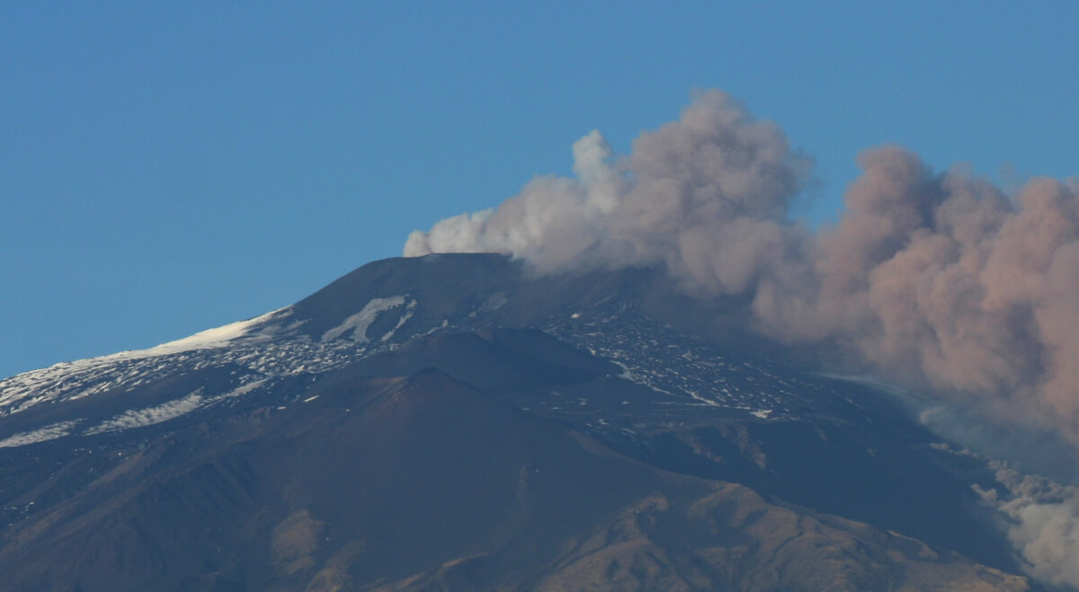 Pennacchio vulcanico grigio e rosato si alza dalla cima innevata dell’Etna e si allunga verso destra sotto un cielo azzurro