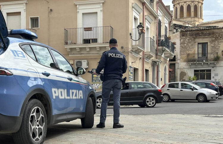 Agente della Polizia di Stato di spalle accanto a un’auto di servizio, in una piazza con palazzi storici e una cupola