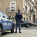 Agente della Polizia di Stato di spalle accanto a un’auto di servizio, in una piazza con palazzi storici e una cupola