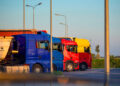 Camion dell’autotrasporto fermi in fila in un piazzale al tramonto, con motrici blu, rosse e gialle