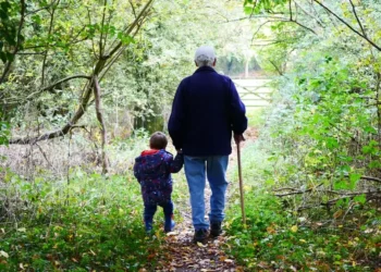 Un nonno con il bastone e un bambino si tengono per mano su un sentiero di bosco coperto di foglie, visti di spalle