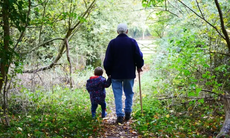Un nonno con il bastone e un bambino si tengono per mano su un sentiero di bosco coperto di foglie, visti di spalle