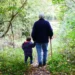 Un nonno con il bastone e un bambino si tengono per mano su un sentiero di bosco coperto di foglie, visti di spalle