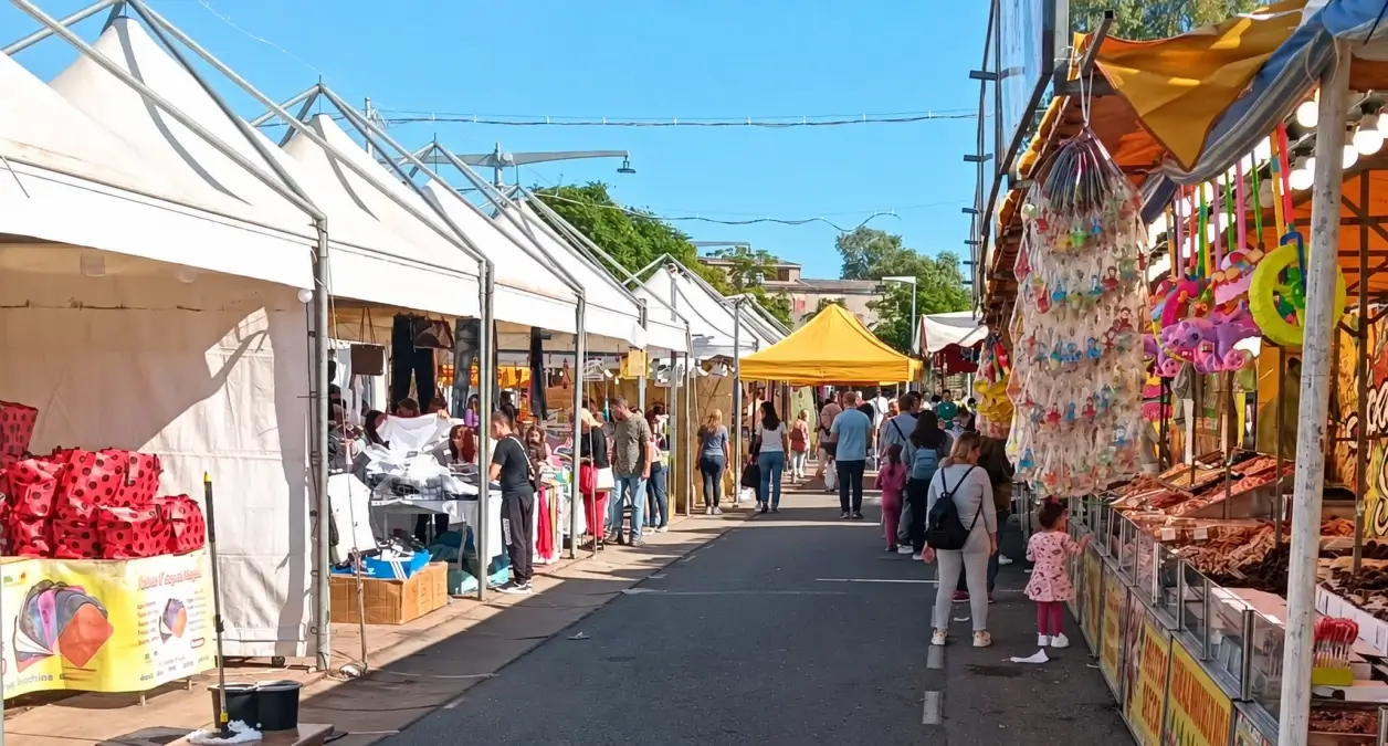 Bancarelle e gazebo bianchi di una fiera dei morti lungo una strada, con visitatori e banchi di giocattoli