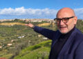 Il geologo Filippo Cappotto con occhiali e giacca blu indica un paese su una collina tra uliveti e prati verdi