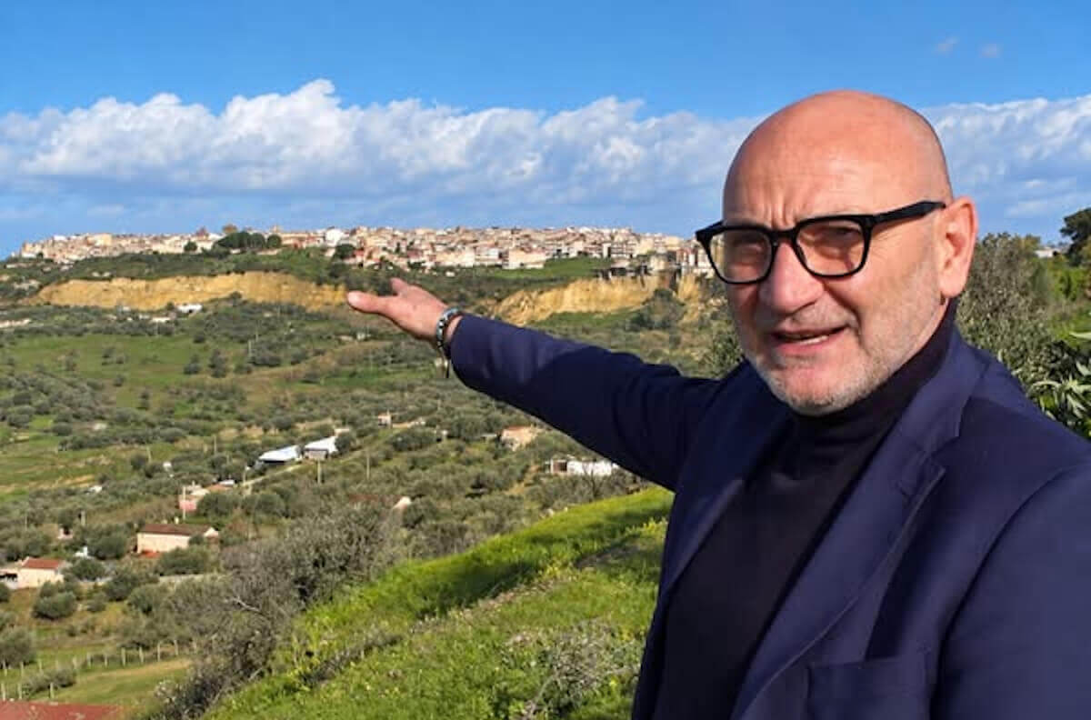 Il geologo Filippo Cappotto con occhiali e giacca blu indica un paese su una collina tra uliveti e prati verdi