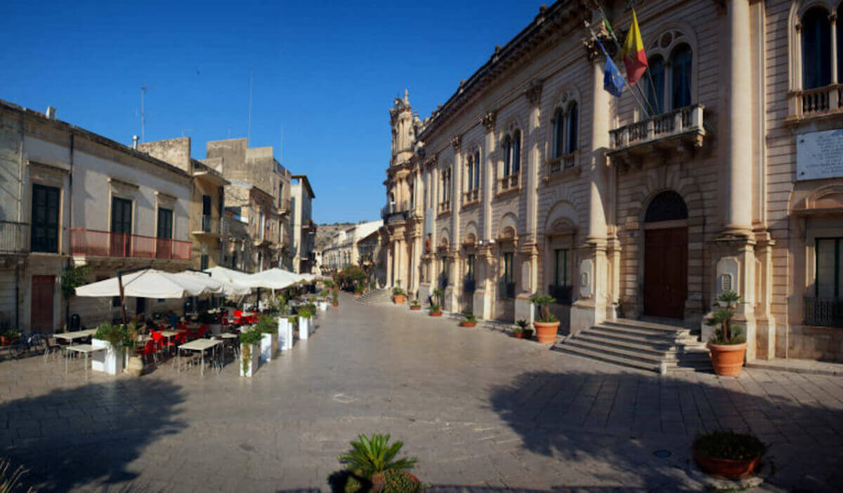 Via Mormino Penna a Scicli con il palazzo del Municipio e i suoi balconi, tavolini all’aperto e una chiesa barocca in fondo
