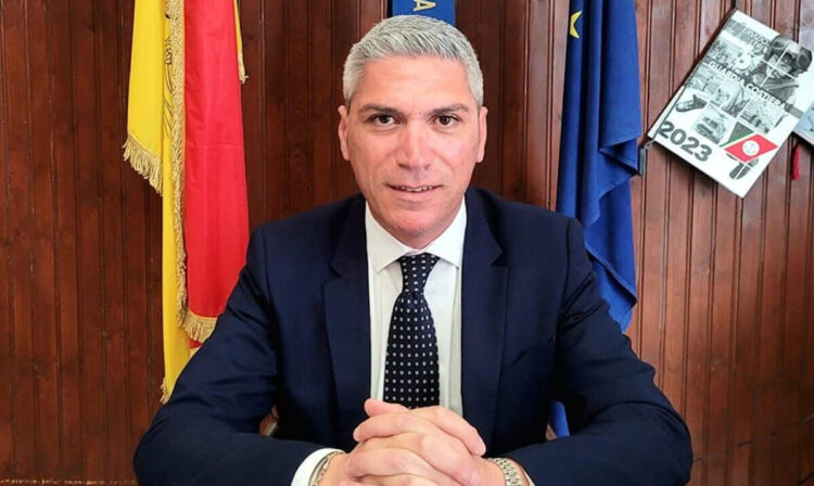 Peppe Dimartino, sindaco di Santa Croce Camerina, seduto alla scrivania con le bandiere di Sicilia, Italia e Unione europea