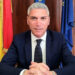 Peppe Dimartino, sindaco di Santa Croce Camerina, seduto alla scrivania con le bandiere di Sicilia, Italia e Unione europea