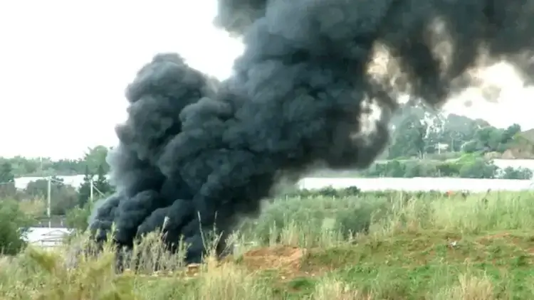 Fumarole di fumo nero denso si alzano dalla vegetazione incolta accanto a una distesa di serre in plastica bianca