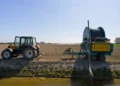 Trattore agricolo arancione fermo accanto a un irrigatore a rullo, su un campo arato lungo un canale d'irrigazione
