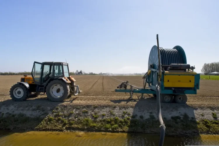 Trattore agricolo arancione fermo accanto a un irrigatore a rullo, su un campo arato lungo un canale d'irrigazione