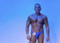 Il bodybuilder Giorgio Buscema in posa sul palco con il numero di gara 002, davanti a uno sfondo azzurro