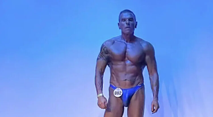 Il bodybuilder Giorgio Buscema in posa sul palco con il numero di gara 002, davanti a uno sfondo azzurro