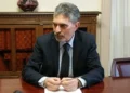 Giovanni Iacono in giacca scura e cravatta, seduto a una scrivania in legno con una penna tra le mani