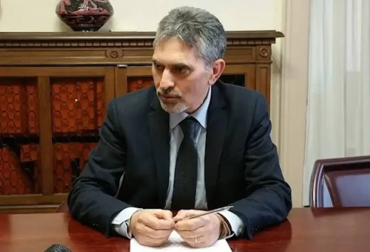 Giovanni Iacono in giacca scura e cravatta, seduto a una scrivania in legno con una penna tra le mani