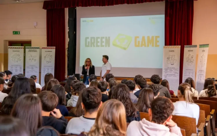 Studenti in un'aula magna seguono la lezione di Green Game, con i pannelli della raccolta differenziata ai lati del palco