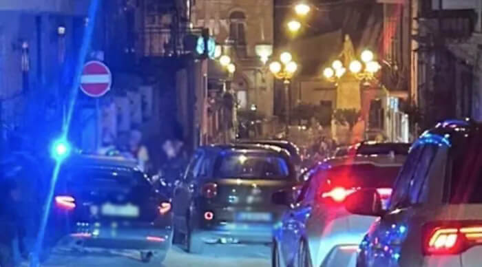 Incidente a Scicli: auto in coda di sera in una via del centro, con un lampeggiante blu e una statua su colonna illuminata