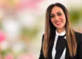 Irene Modica sorridente, capelli lunghi castani, giacca nera e camicia bianca con fiocco, su sfondo floreale sfocato