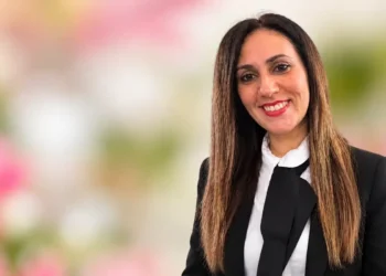 Irene Modica sorridente, capelli lunghi castani, giacca nera e camicia bianca con fiocco, su sfondo floreale sfocato