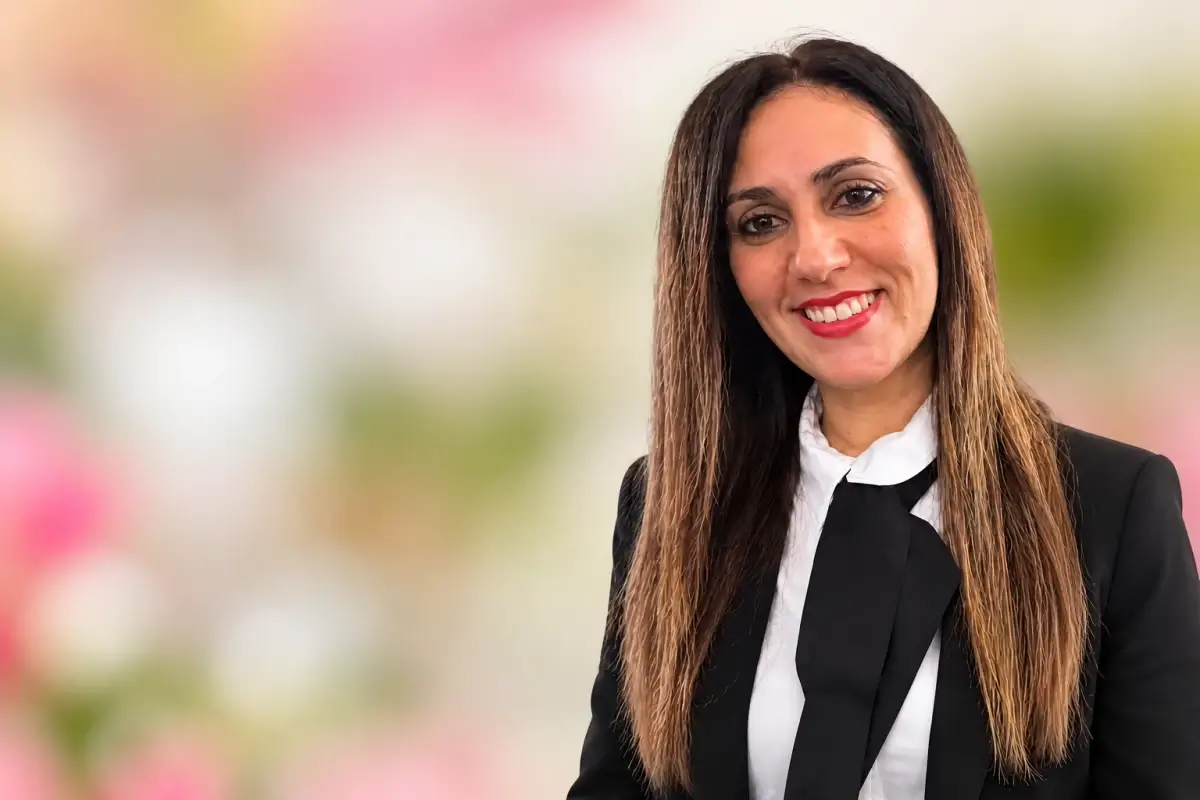 Irene Modica sorridente, capelli lunghi castani, giacca nera e camicia bianca con fiocco, su sfondo floreale sfocato