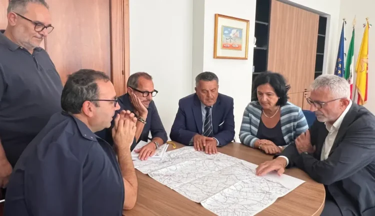 Sei persone attorno a un tavolo esaminano una mappa delle strade regionali tra Ragusa e Scicli in un ufficio con bandiere