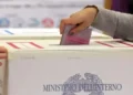 Una mano inserisce una scheda elettorale rosa in un’urna di cartone con il logo del Ministero dell’Interno