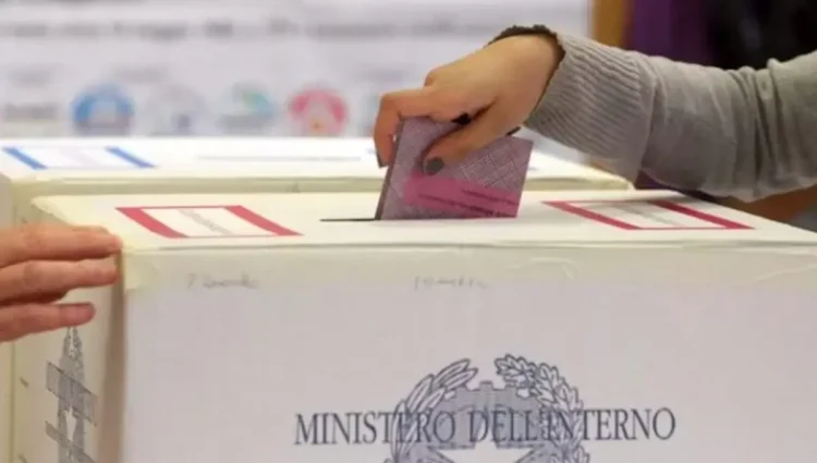 Una mano inserisce una scheda elettorale rosa in un’urna di cartone con il logo del Ministero dell’Interno