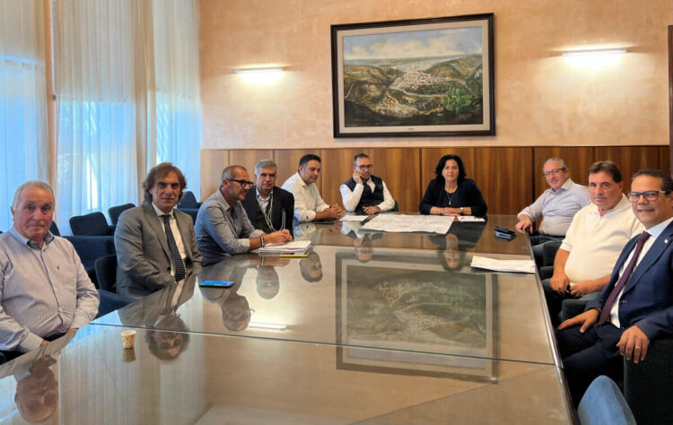 Riunione sui lavori della SR 100 per la Pantano-Geraldo: undici persone attorno a un tavolo di vetro con planimetrie stradali