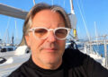 Luigi Giuga, l’ottico di Rosolini, con occhiali bianchi e maglia nera, seduto in barca a vela in un porto turistico
