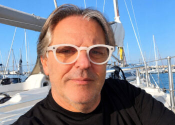 Luigi Giuga, l’ottico di Rosolini, con occhiali bianchi e maglia nera, seduto in barca a vela in un porto turistico