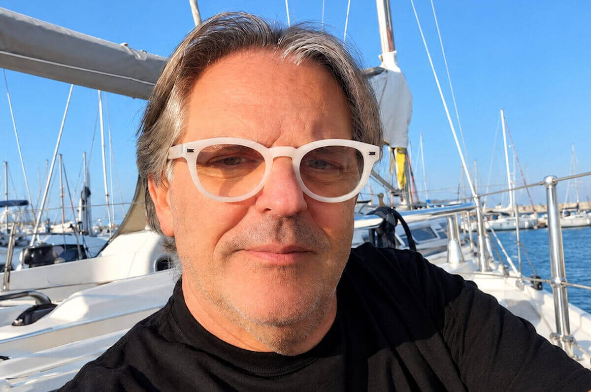 Luigi Giuga, l’ottico di Rosolini, con occhiali bianchi e maglia nera, seduto in barca a vela in un porto turistico