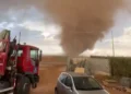 Tromba d’aria carica di polvere su una campagna durante il maltempo, vista da un cortile con un camion con gru e un’auto grigia
