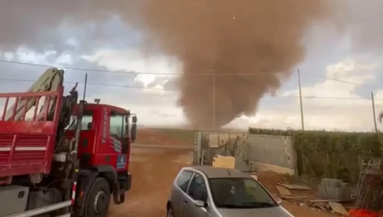 Tromba d’aria carica di polvere su una campagna durante il maltempo, vista da un cortile con un camion con gru e un’auto grigia