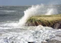 Onde di maltempo si infrangono su un costone erboso, con schiuma bianca sulla riva e mare agitato sotto un cielo grigio