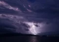 Fulmine durante un temporale di maltempo sul mare di notte, tra nuvole scure e una costa illuminata