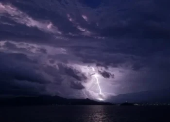 Fulmine durante un temporale di maltempo sul mare di notte, tra nuvole scure e una costa illuminata