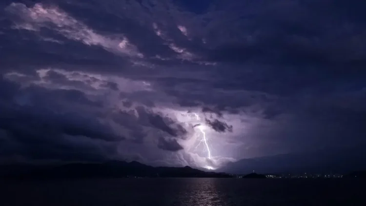 Fulmine durante un temporale di maltempo sul mare di notte, tra nuvole scure e una costa illuminata