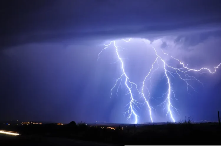Fulmini durante un temporale notturno di maltempo, sotto un cielo viola, con le luci di un centro abitato all’orizzonte
