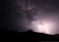 Due fulmini squarciano il cielo notturno durante un temporale, sopra una collina alberata in controluce: scena di maltempo