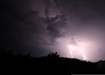 Due fulmini squarciano il cielo notturno durante un temporale, sopra una collina alberata in controluce: scena di maltempo