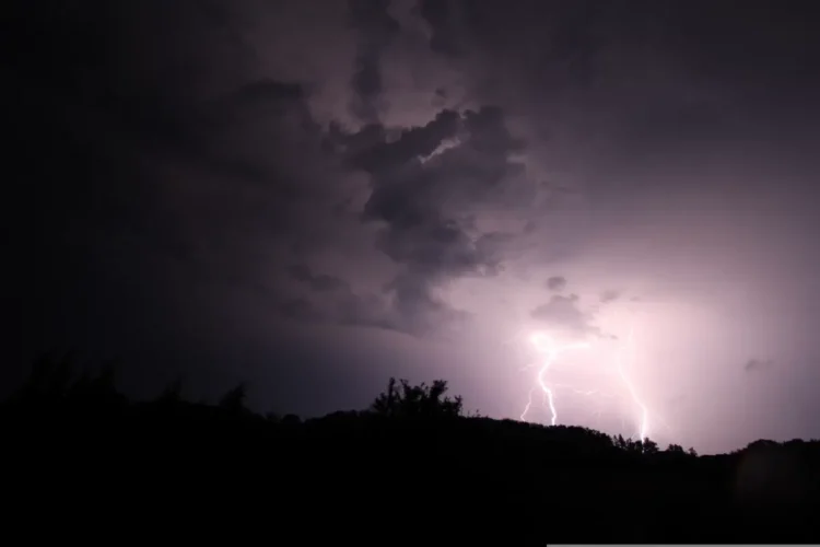 Due fulmini squarciano il cielo notturno durante un temporale, sopra una collina alberata in controluce: scena di maltempo