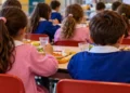 Bambini di spalle con grembiuli rosa e blu pranzano ai tavoli di una mensa scolastica. Immagine generata con intelligenza artificiale